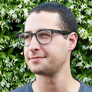A light-skinned man with short dark hair, black square glasses, and light stubble, wearing a blue tshirt and standing in three-quarter profile against a background of bush with white flowers.