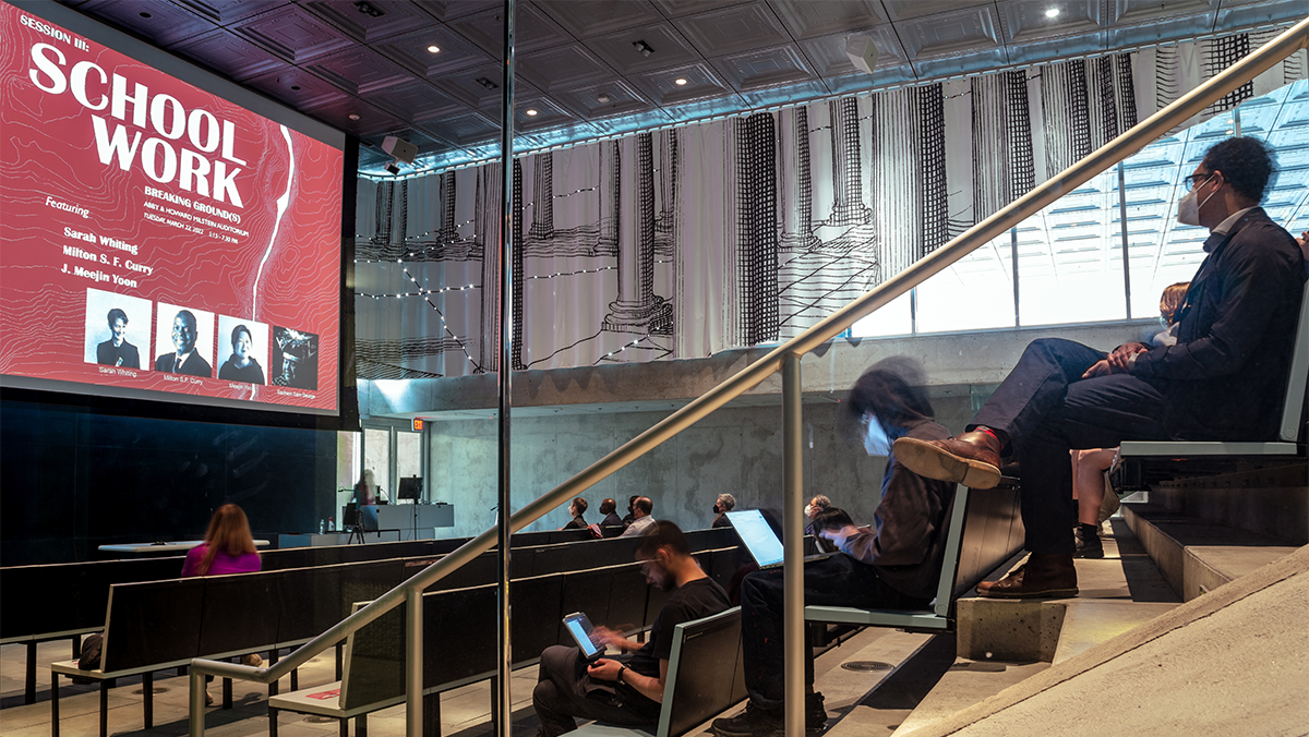 Red image with white text on a screen in an auditorium with an audience