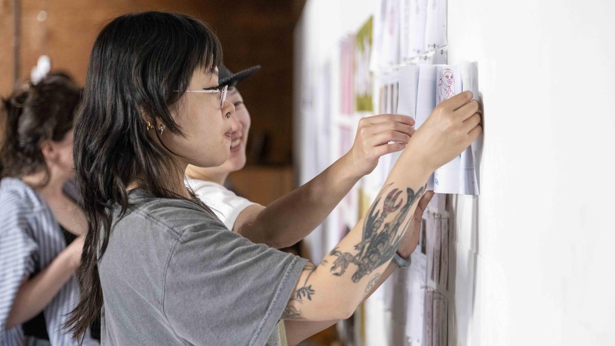 Person with long dark hair in a grey shirt with tattoos hanging papers on a white wall.