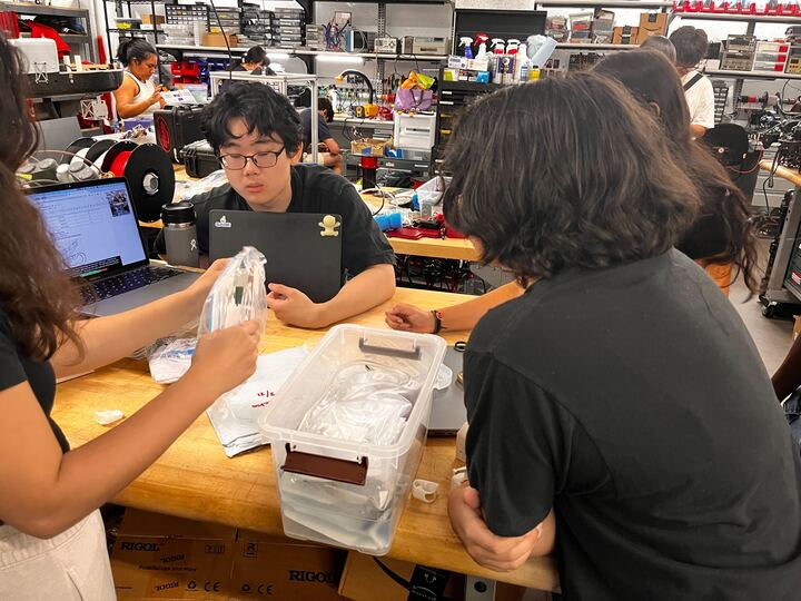 students collaborate around a workshop table, examining clear plastic parts in a busy makerspace.