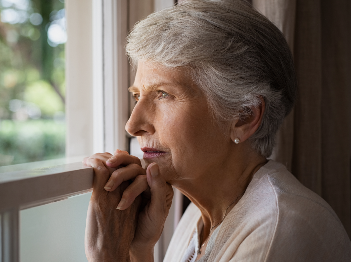 An older woman with short gray hair gazes thoughtfully out a window, resting her hands lightly on the windowsill as soft daylight illuminates her face.