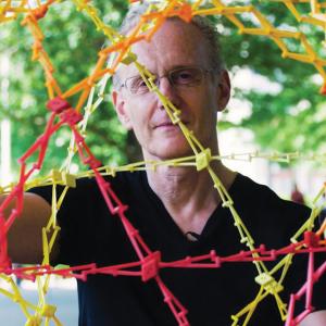 A senior white man wearing glasses and a black short looking at the camera and stretching a colorful circular apparatus with both hands.