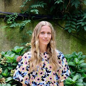 A blonde woman in a colorful top standing in front of shrubbery