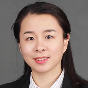 A woman with tied-back dark hair, wearing a black blazer and a light striped shirt, smiling softly against a plain gray background.