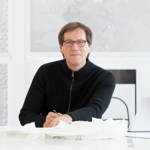 A portrait of a white man with glasses and short hair whereing a black zip up sweater.