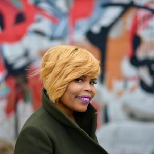 A person with short blonde hair and purple lipstick wearing a green coat standing in front of a multicolored art wall.