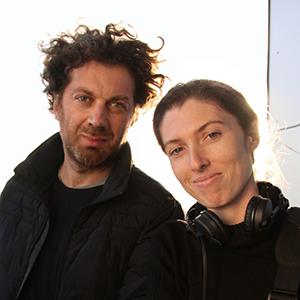 A man and a woman wearing dark clothing, standing adjacent to one another. 