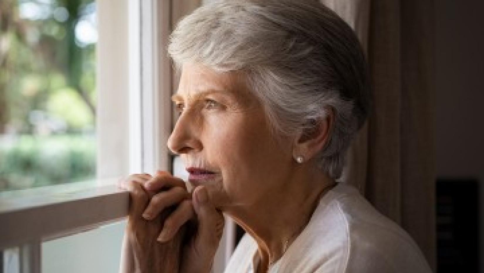 An older woman with short gray hair gazes thoughtfully out a window, resting her hands lightly on the windowsill as soft daylight illuminates her face.