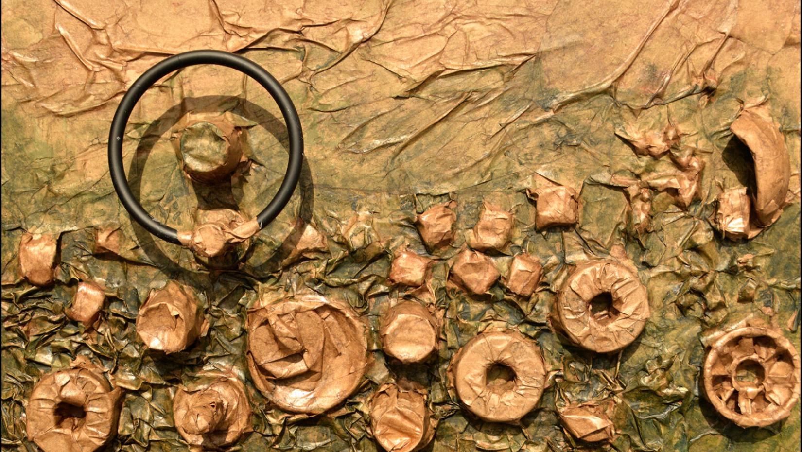Abstract mixed-media relief with copper-toned circular forms and a black ring embedded in a wrinkled green surface of reclaimed materials.