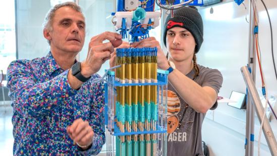 A professor and student collaborate in a bright lab, assembling a tall blue-and-gold mechanical structure as part of a hands-on engineering project.