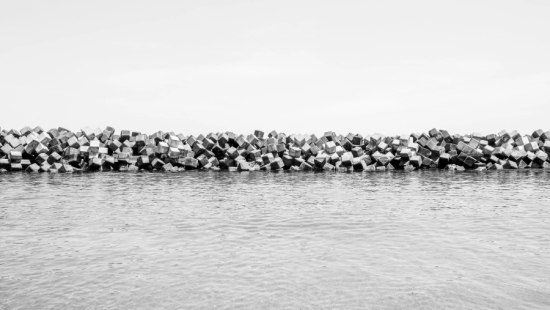 Black-and-white photograph of stacked concrete breakwater blocks forming a low barrier along calm water beneath an open, overcast sky.