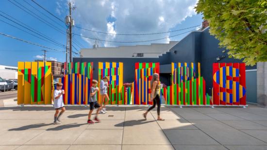 People walk past Apart, Together, a colorful installation of angled red, yellow, green, and blue panels that form shifting patterns and the word “YES” along a Columbus, Indiana sidewalk.
