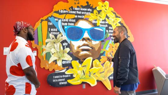 Two men stand in conversation before a vibrant mural of a young Black girl wearing oversized blue sunglasses, surrounded by yellow lilies and overlaid with text from Toni Morrison’s writings, installed on a red wall in Cornell’s Morrison Hall.