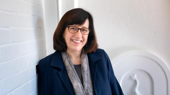 A woman with glasses and shoulder length brown hair wearing a blue jacket with a grey scarf.
