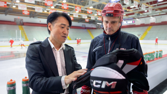 Two men in the stands of a hockey rink holding protective gear and chatting