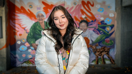 A person with a white coat standing in front of a bright colorful painted mural of two individuals.