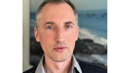 A man wearing a collared shirt with a jacket standing in front of an ocean image.