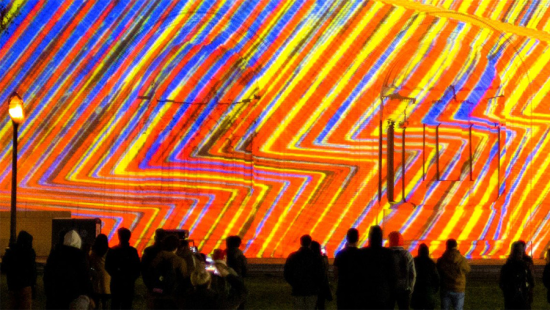 Blue, orange, and yellow zig-zag lines of light projected onto the side of a building at night with spectators in front