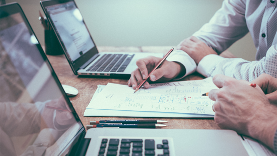 Desktop with two laptops, pens, and two pairs of hands working on a stack of paper positioned in between