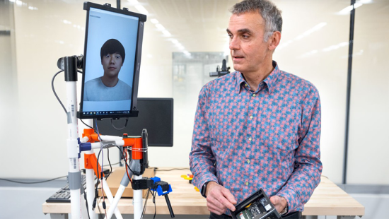 Man wearing a bright button-down shirt standing next to a tablet displaying a rendered student face that is mounted on a stand 