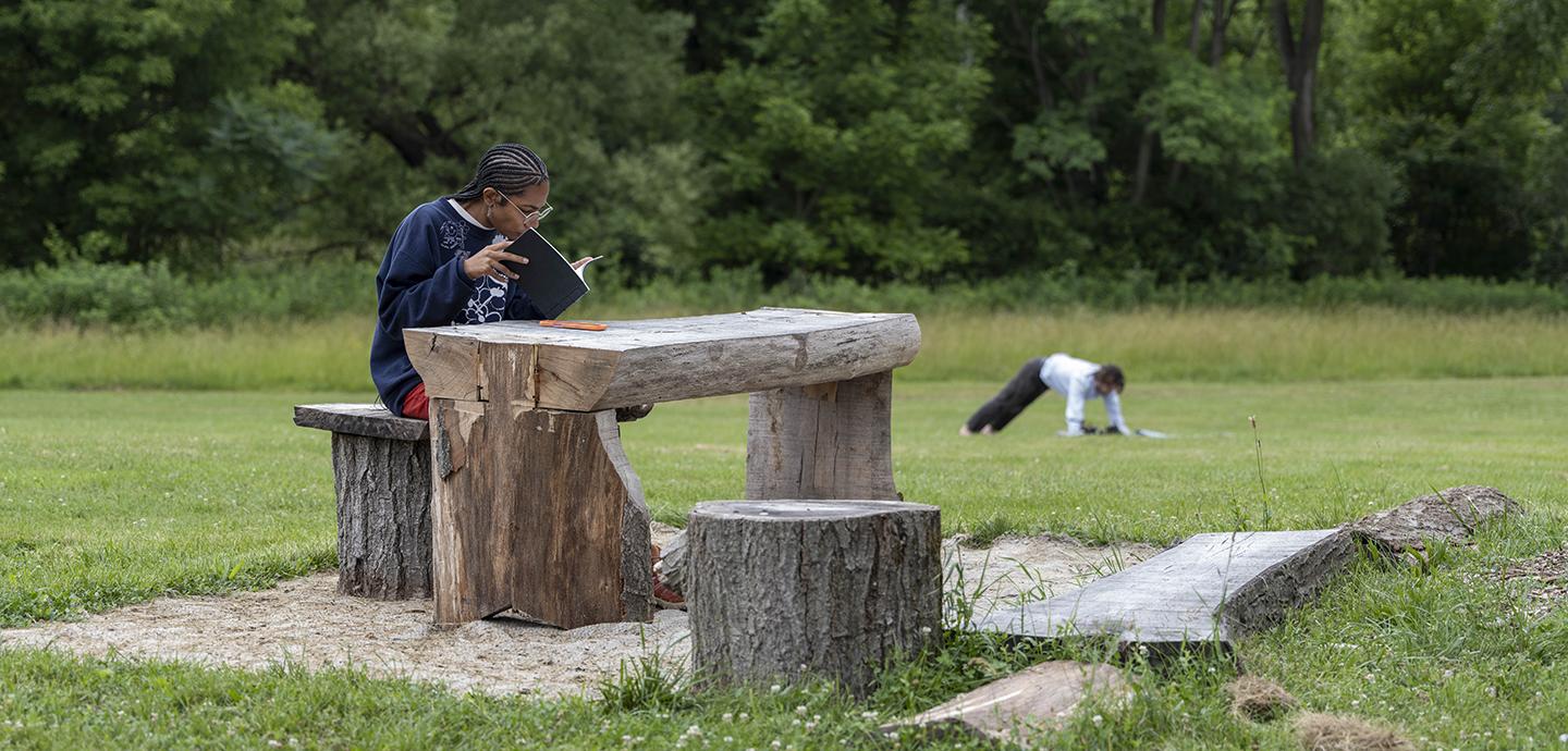 Person sitting on a wooden log at a wooden carved table outside with green trees in the background.