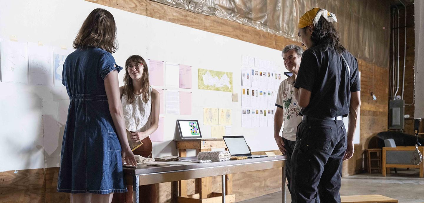 Four people gathered around a small table with 2 screens and various papers pinned behind on a white work board.