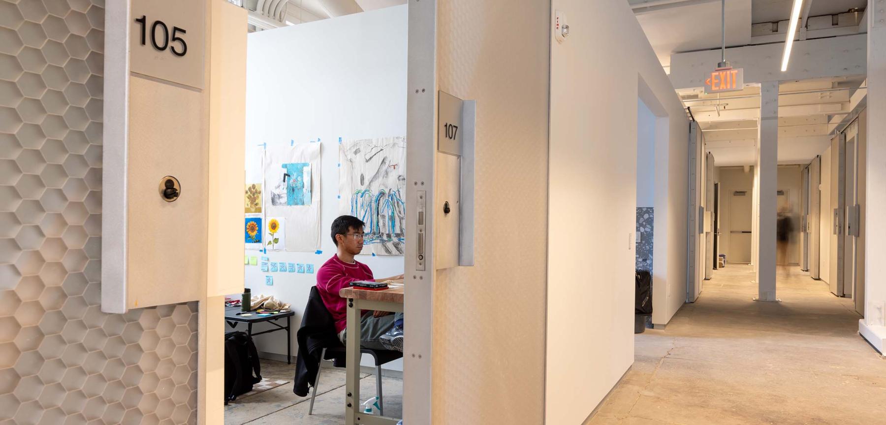 Person wearing red shirt sitting at a desk with drawings on the wall looking down a long hallway.