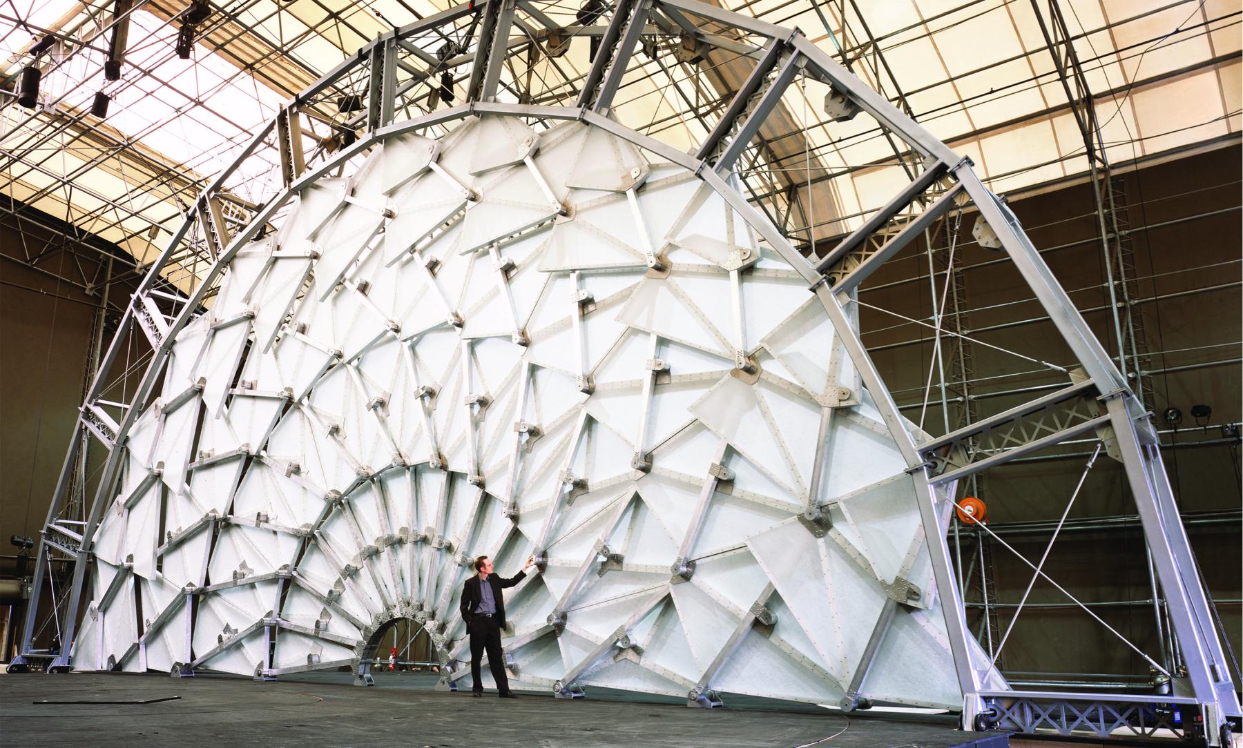 A person stands in front of a large structure in the shape of a semi-circle made by multiple articulated geometric pieces.