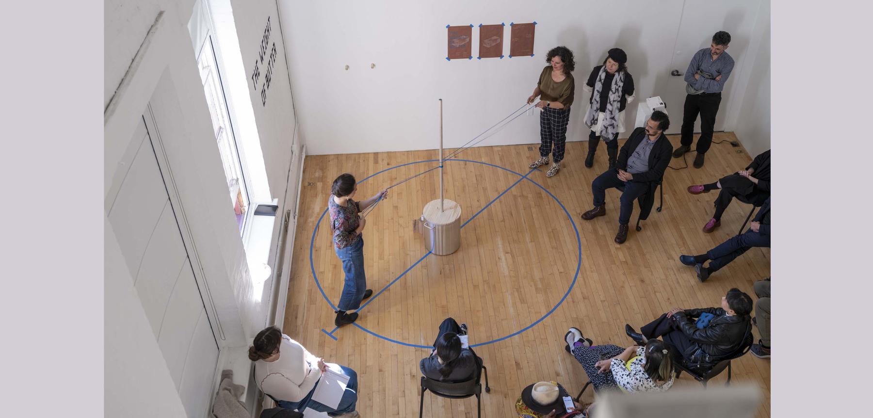 Two people holding pieces of string around a blue circle on the floor with others gathered around.