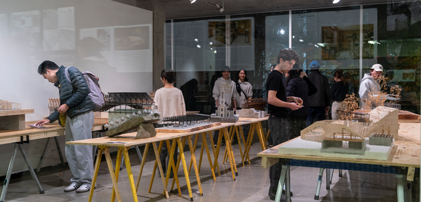 People gather around tables in a gallery covered with wooden and concrete architectural models