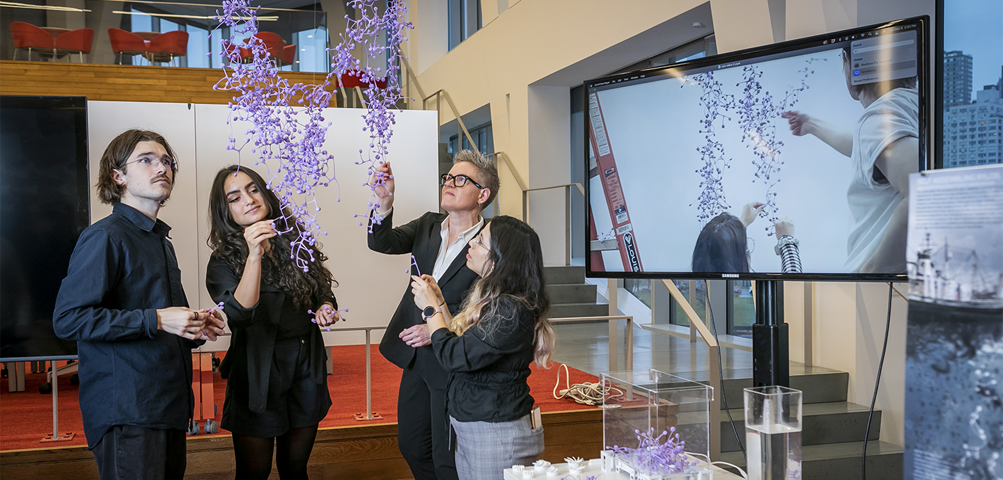 Four people looking at a hanging sculpture made of small purple objects.