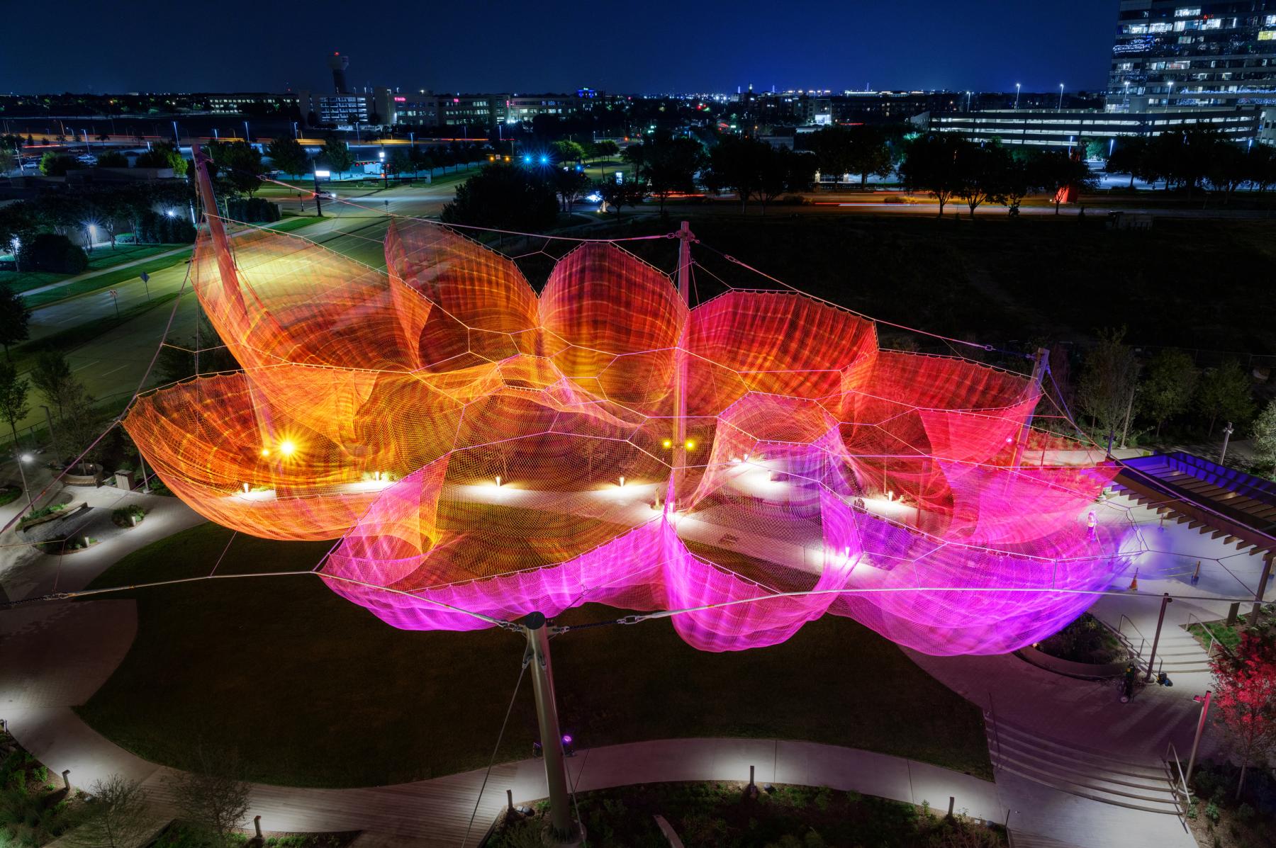 A big sculpture that resembles an open flower made of fabric hanging above an open park and illuminated with coloful lights in the night.
