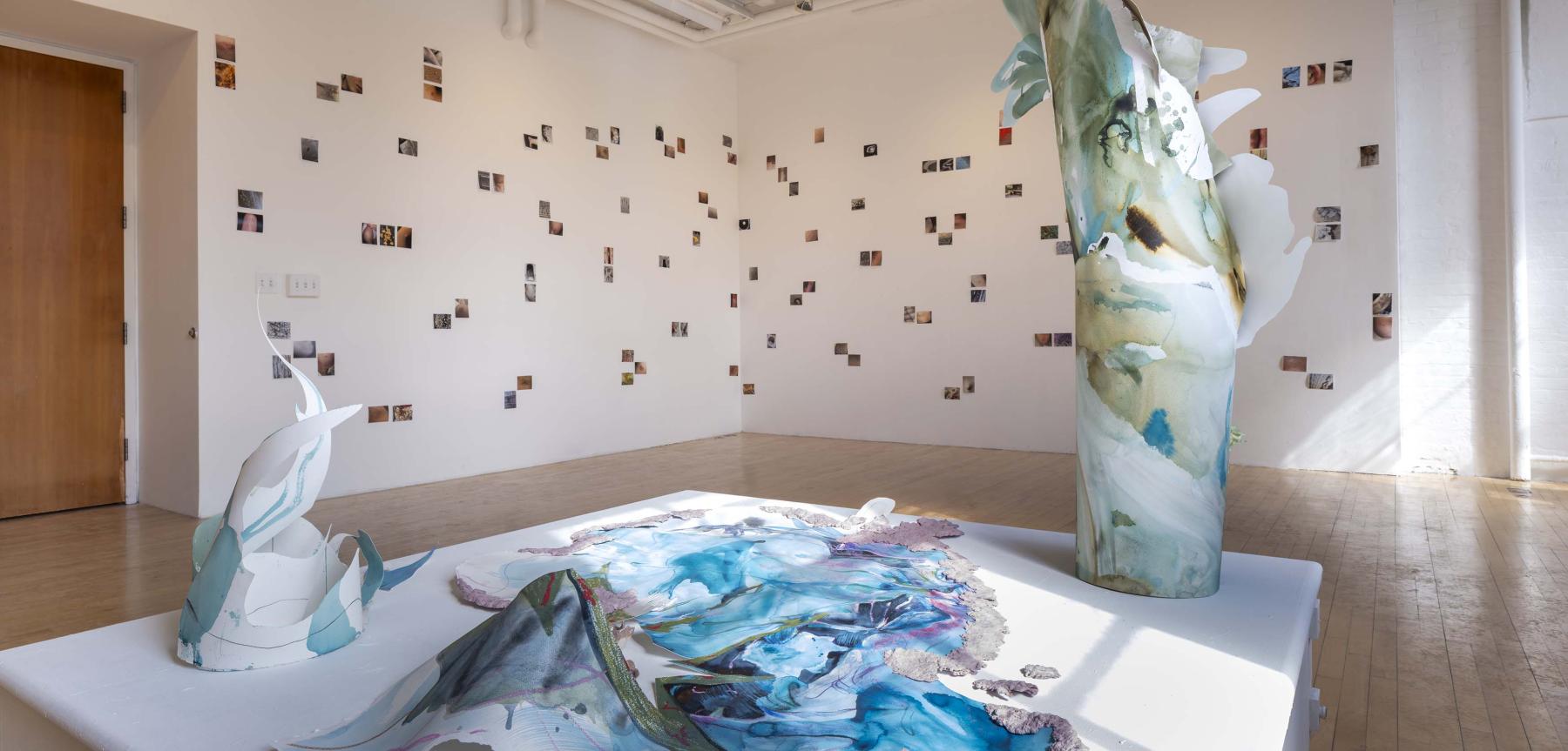 Various abstract water color paper sculptures on a white pedestal in front of small pictures hung on a a white wall.