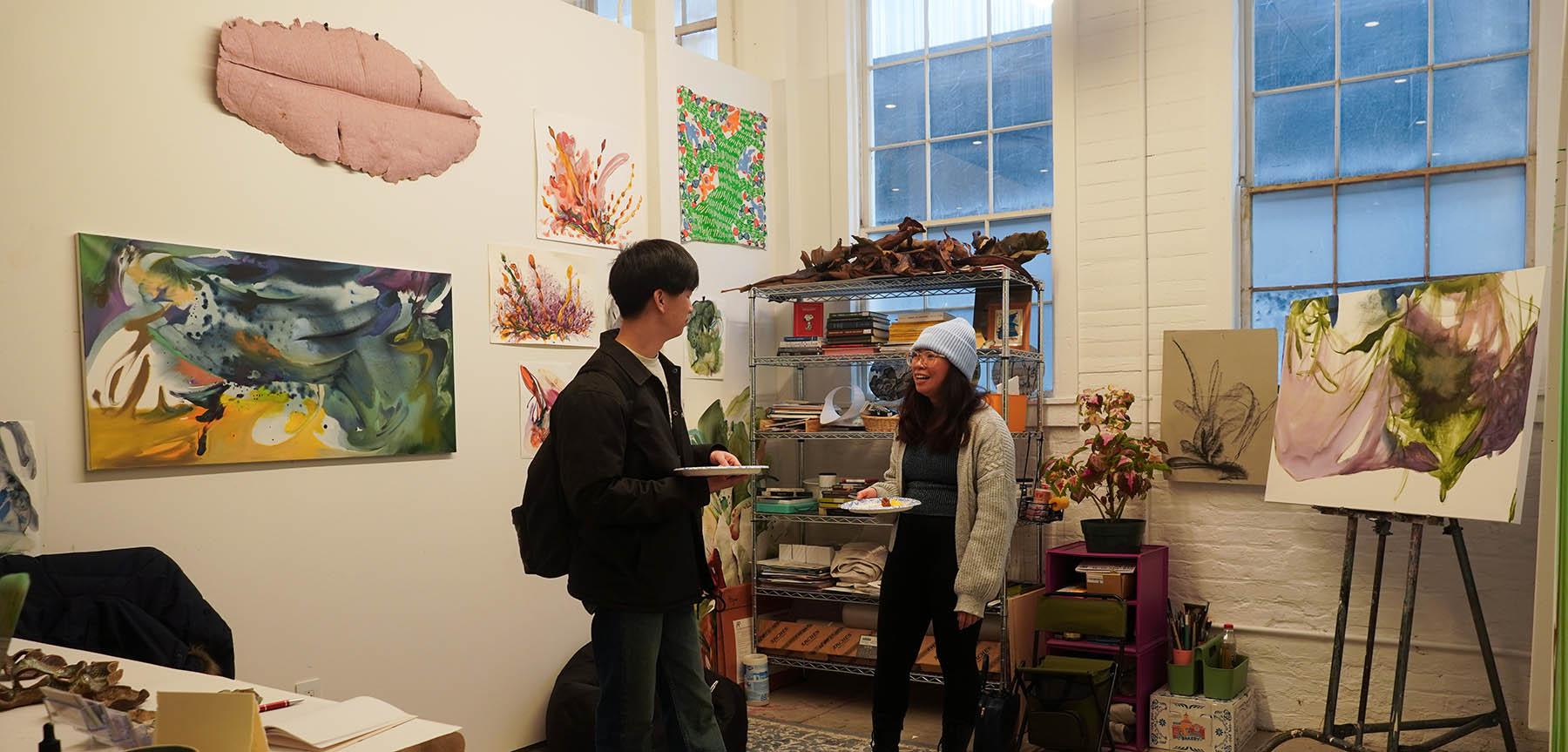 Two people in a room with various paintings and drawings on a wall. 