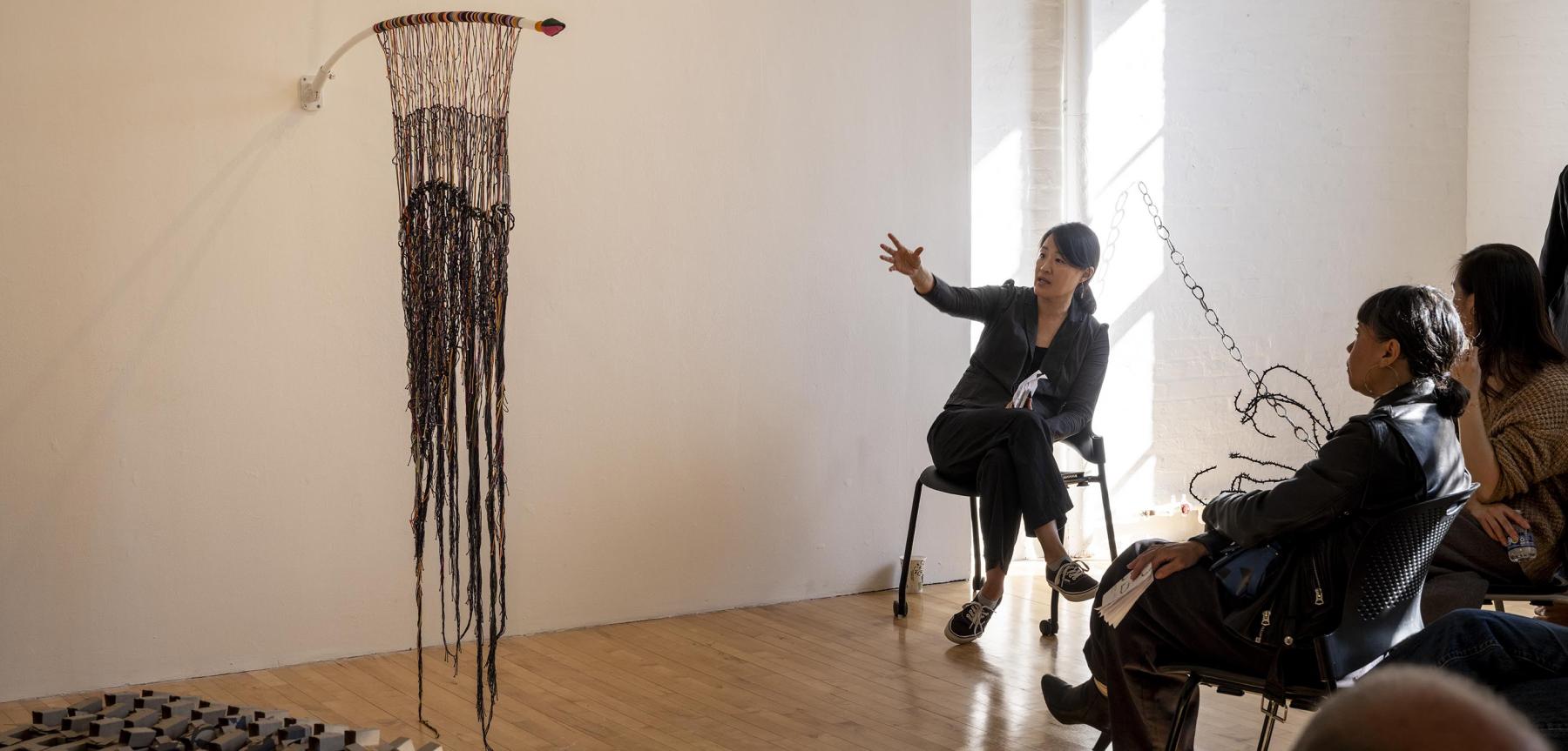 People wearing dark clothing sitting down discussing a hanging yarn fabric piece.