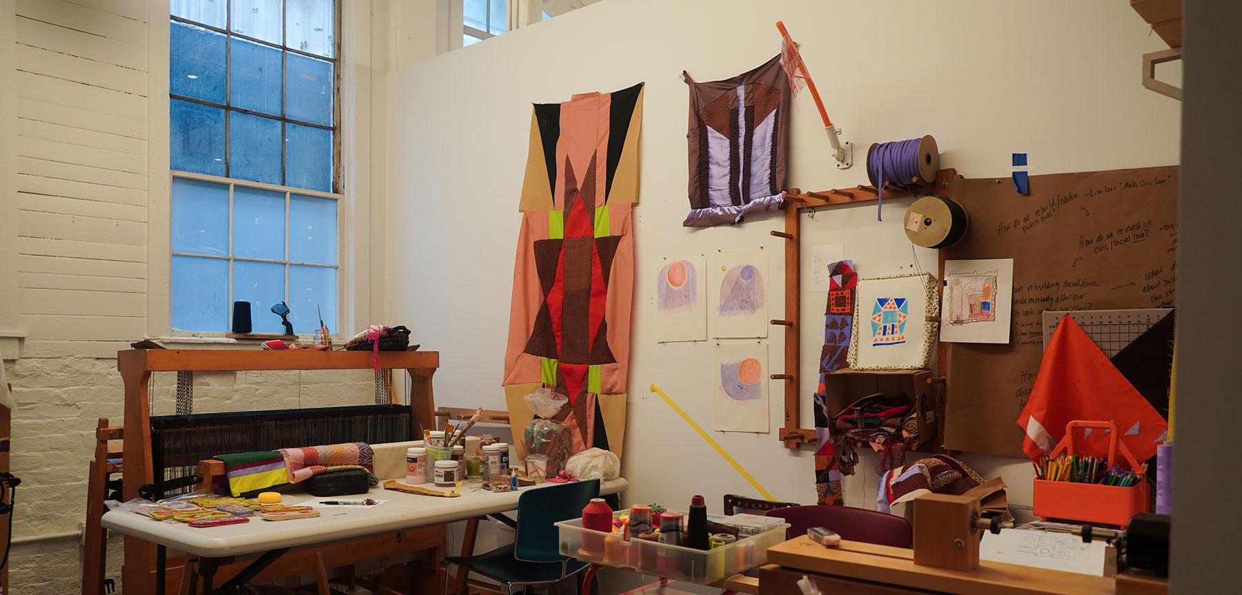 Room with various fabric pieces hanging on a wall with a loom and other sewing materials in the space.