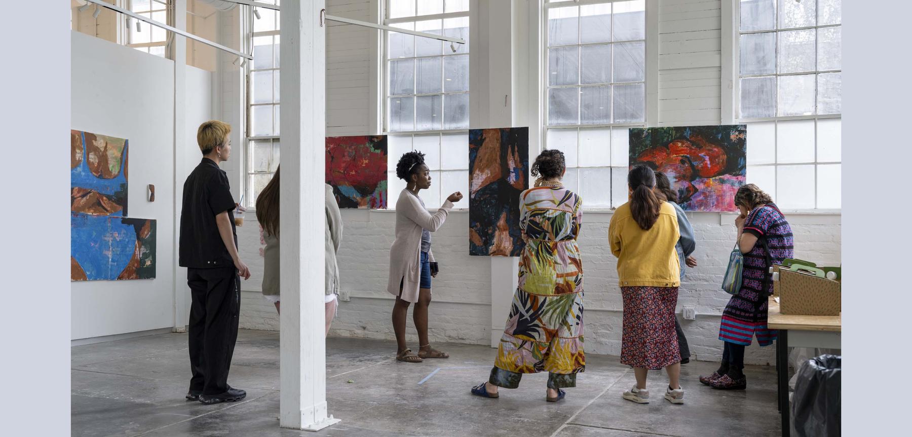 Seven people facing various paintings on display in a bright white and gray room.