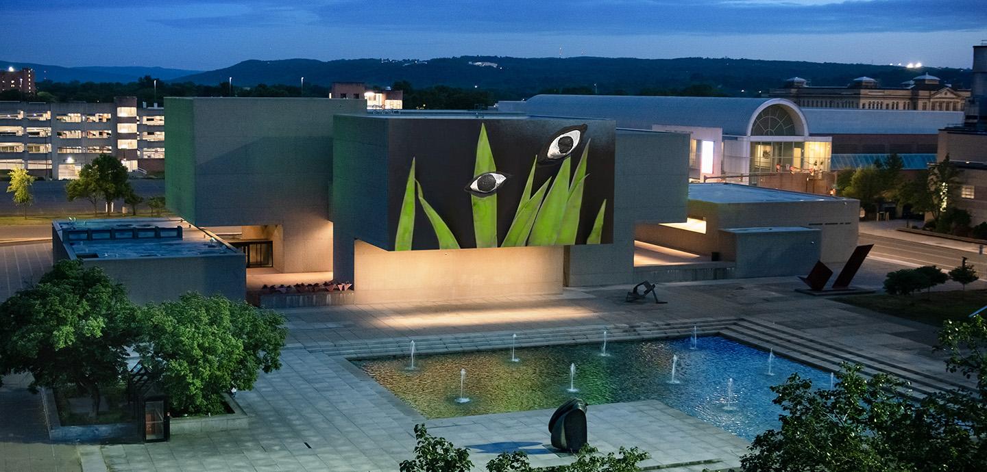 A view of a stone courtyard with an L-shaped pool and numerous small fountains. Behind it is a building painted with blades of grass and two large eyes.