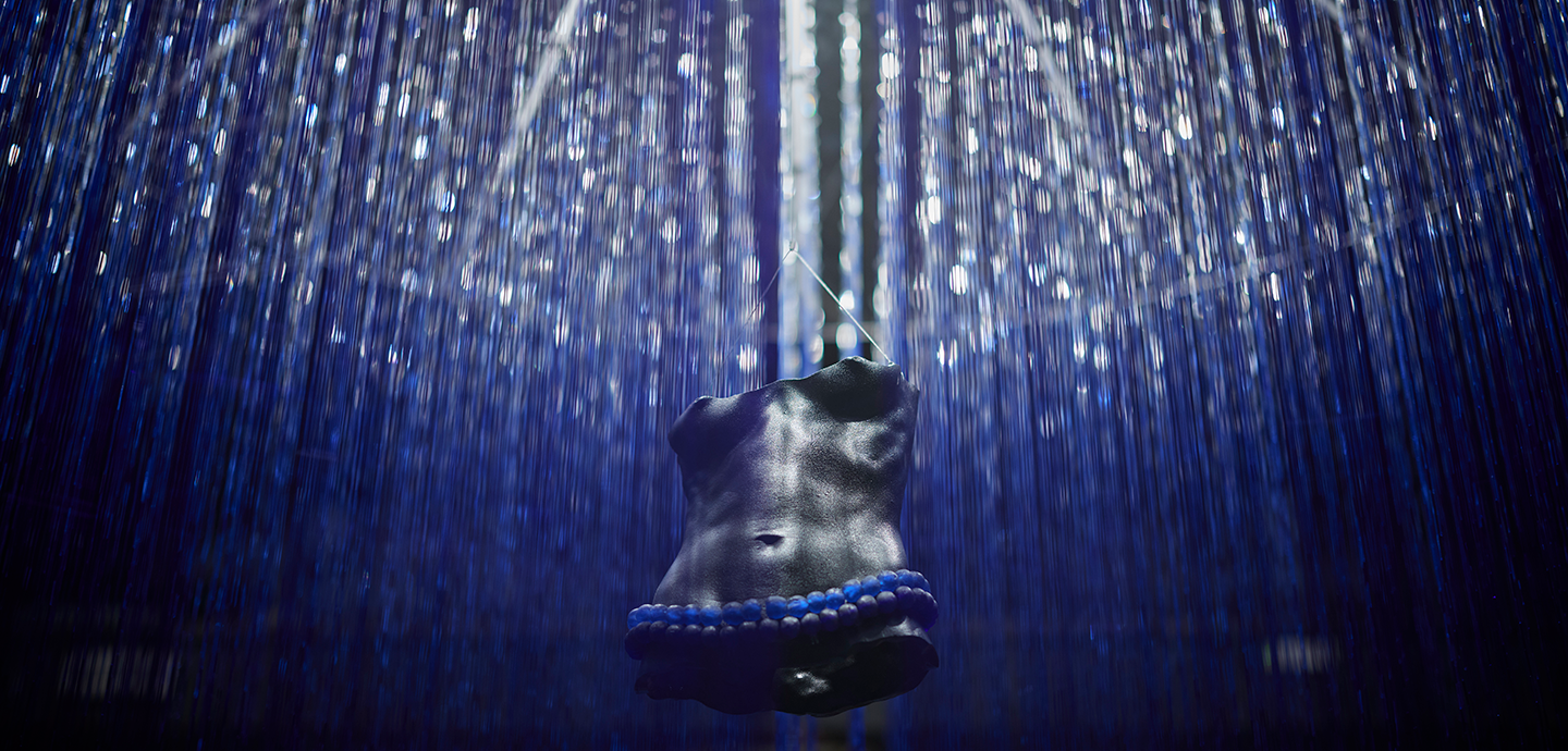 Sculpture of a dark-skinned chiseled feminine torso with beads around its waist is surrounded by clear monofilament, reminiscent of rain.