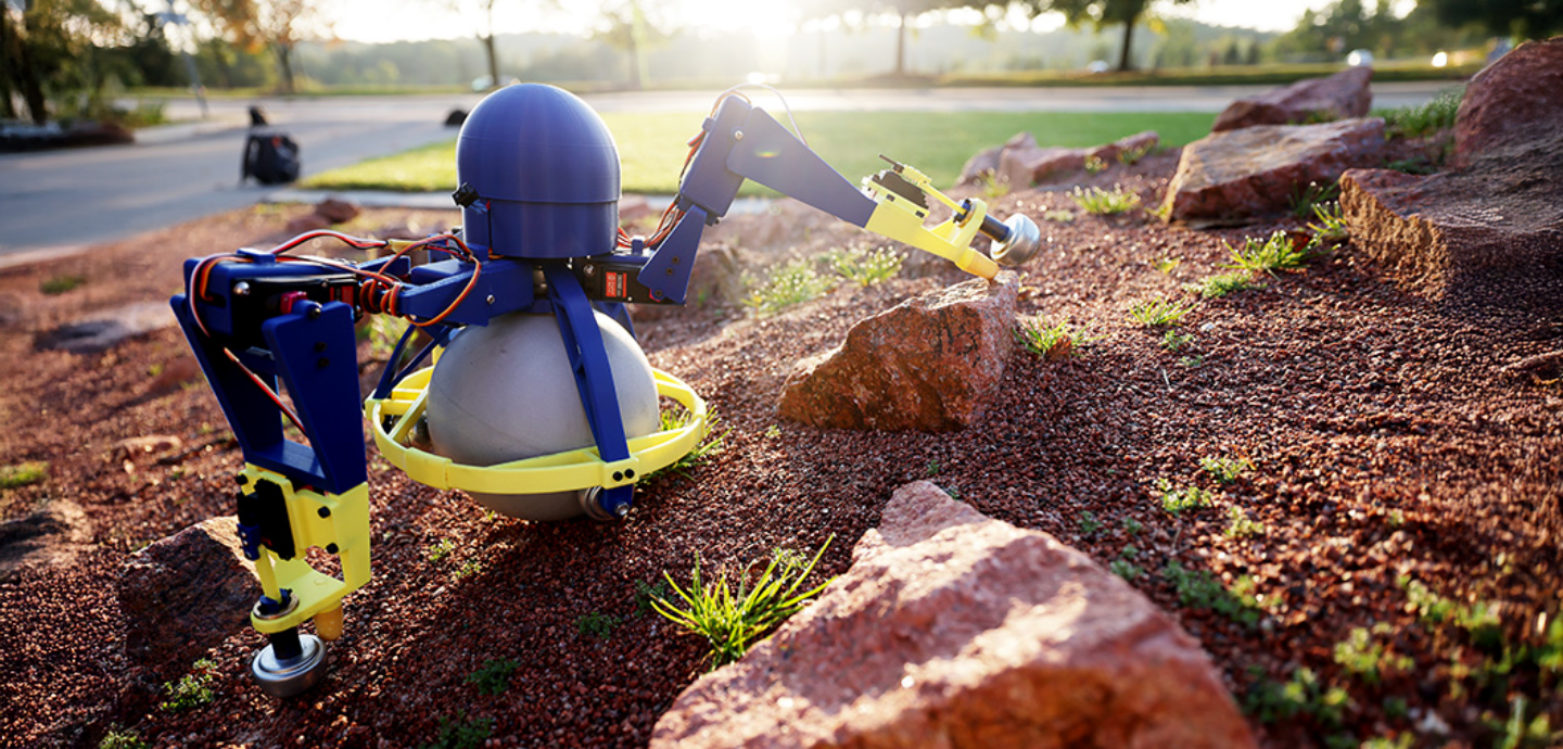 A mechanism with robotic arms made of metal and plastic parts crawling among a rocky field.