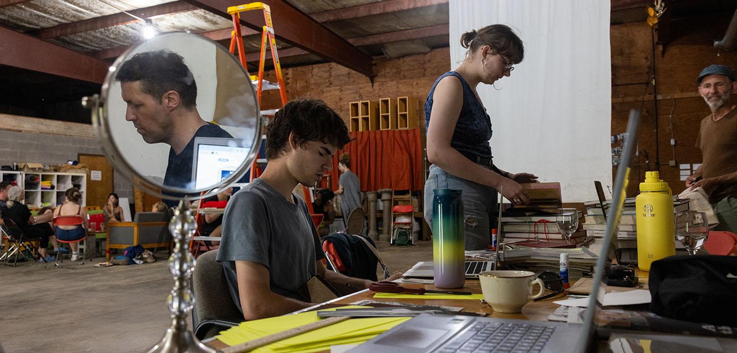 People working on laptops, reading books, in discussion in a large workshop area.