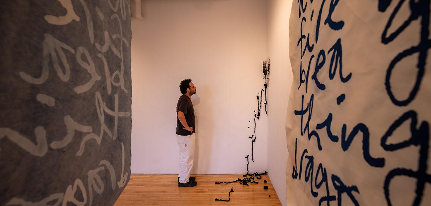 Man wearing dark shirt and white pants looking at abstract dark colored fabric and thread hanging from a white wall.
