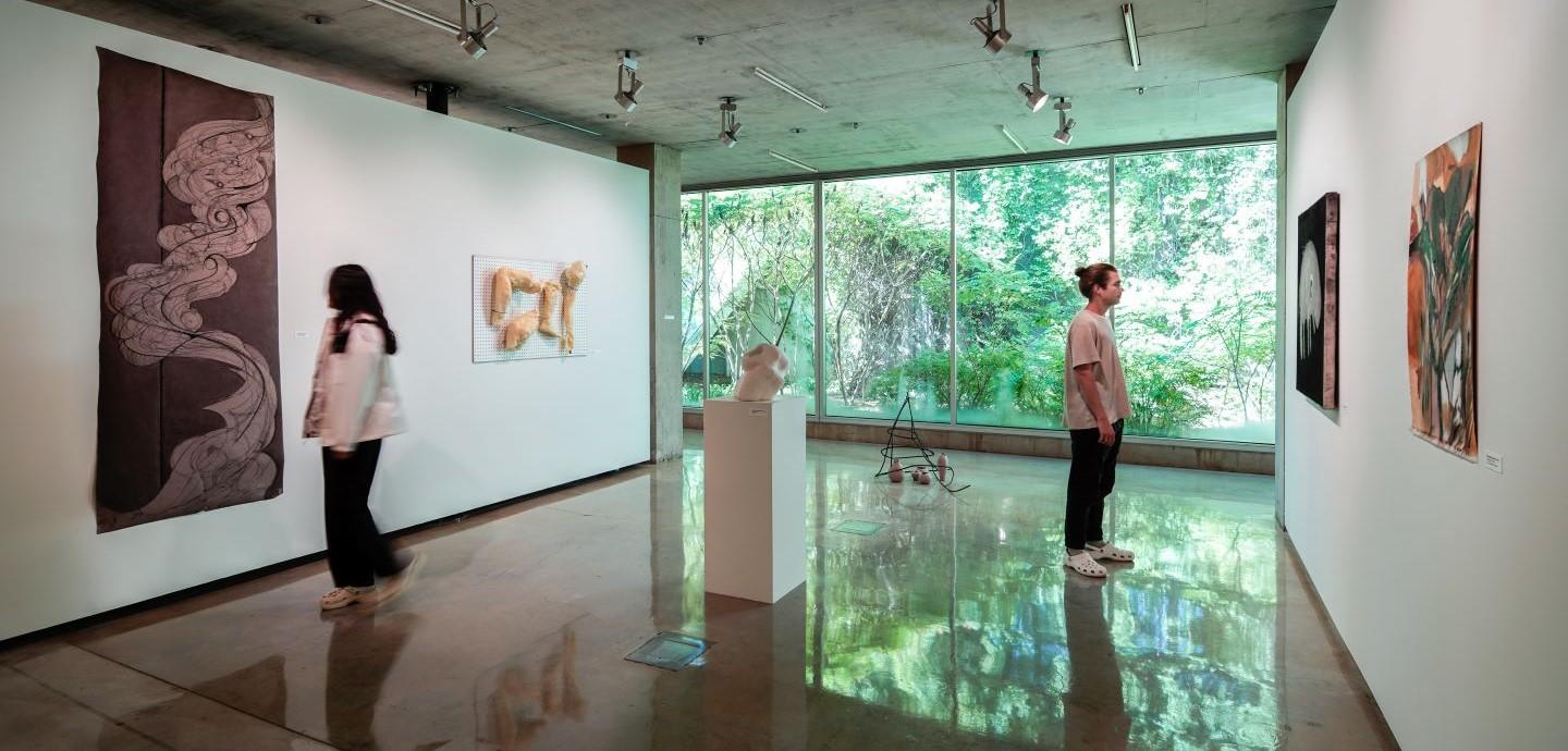 Guests look at student artwork in a bright gallery space bathed in reflective light, a glass wall looking outdoors in the background