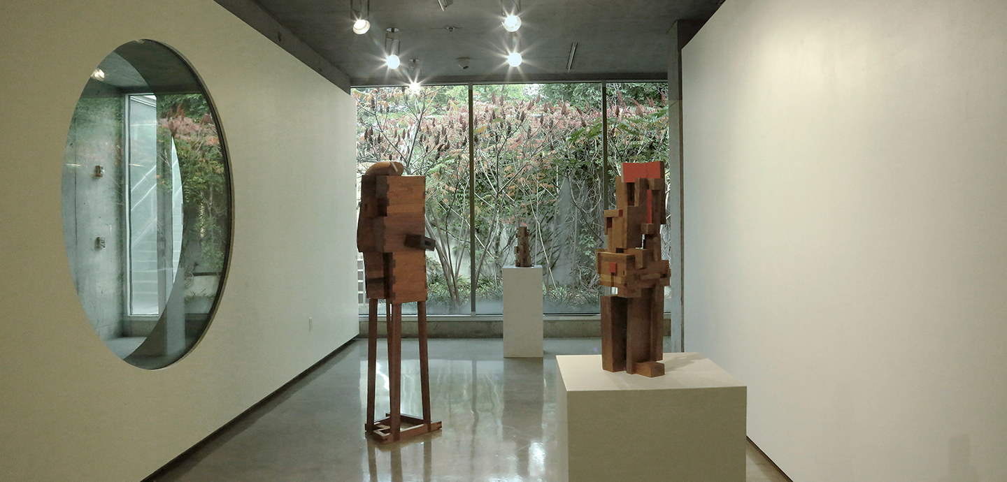 Abstract wooden sculpture piece with various sized rectangular pieces put together displayed on white pedestals. 
