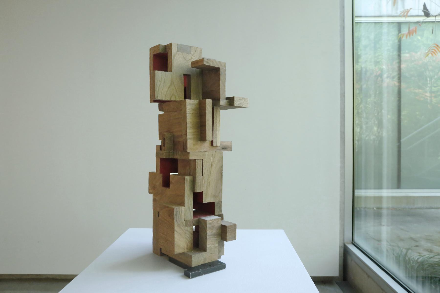 Abstract wooden sculpture piece with various sized rectangular pieces put together displayed on a white pedestal. 