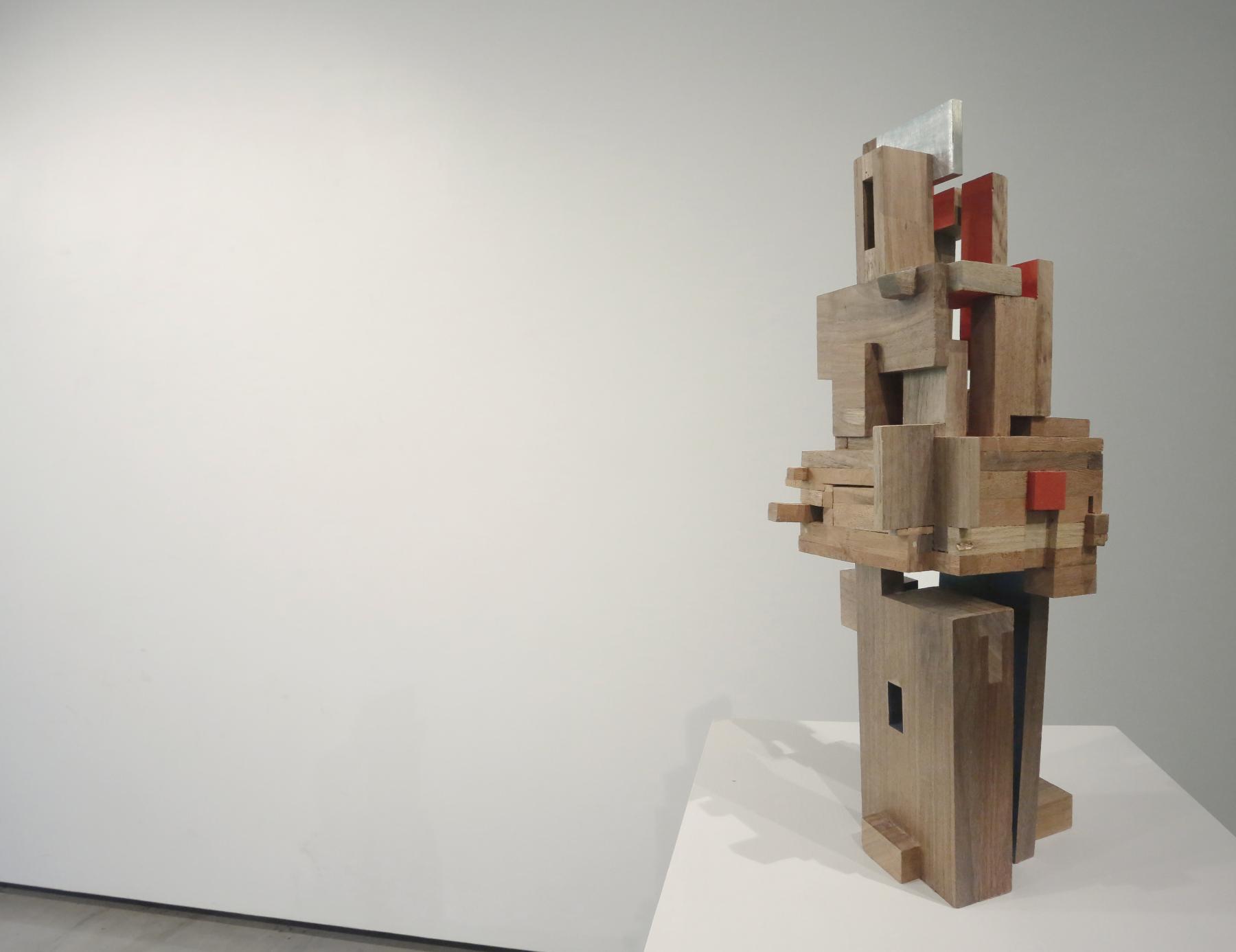 Abstract wooden sculpture piece with various sized rectangular pieces put together displayed on a white pedestal. 