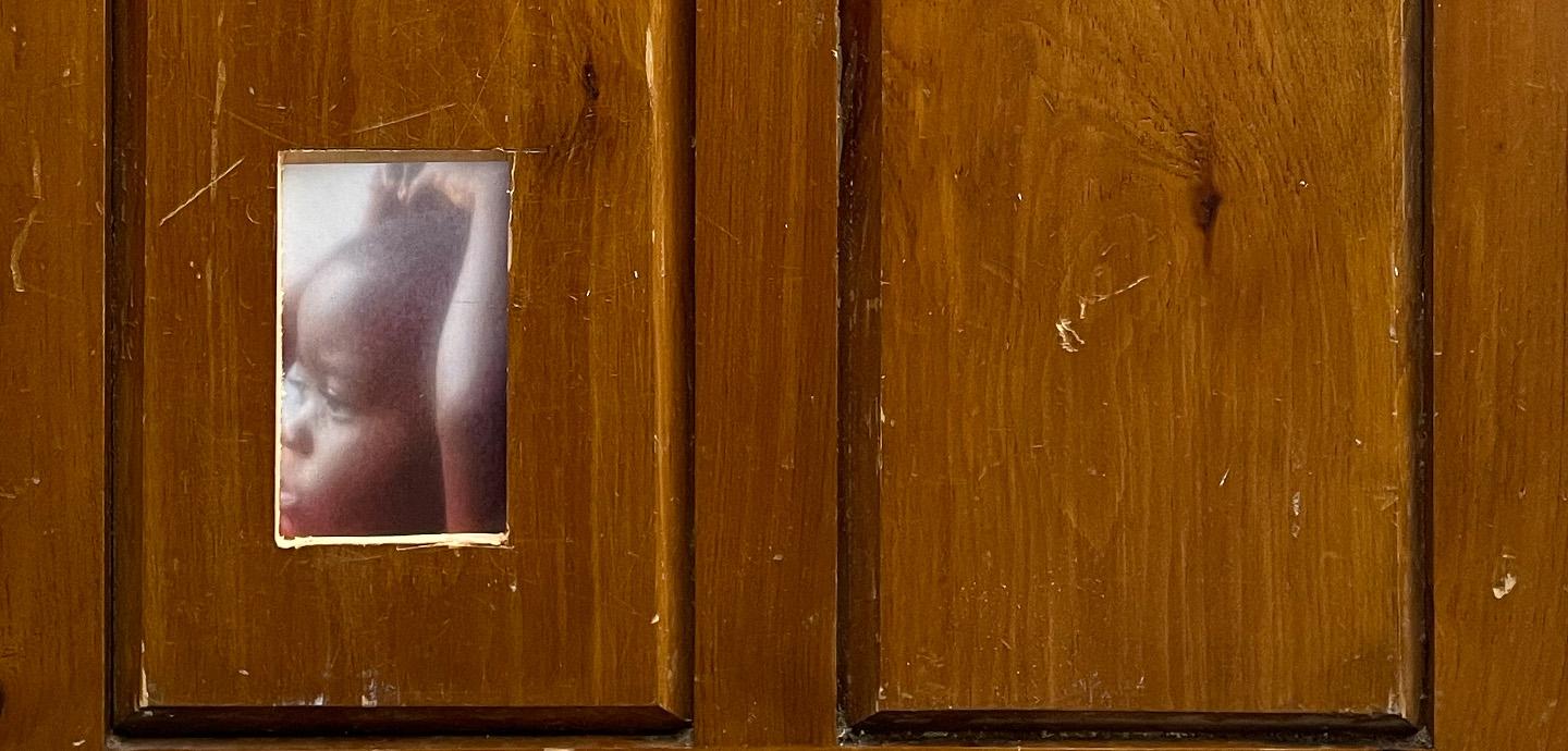 A wood panel with a small rectangular cutout, revealing a photo of a small child with their hands on their head.