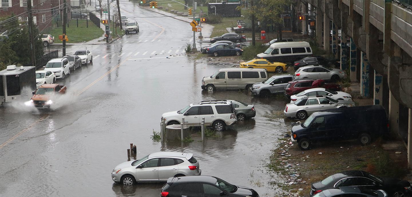 A rainy city street is flooded. On the left, a red pickup truck sprays up water as it travels down the street. A dozen cars are parked on the right next to a subway overpass.