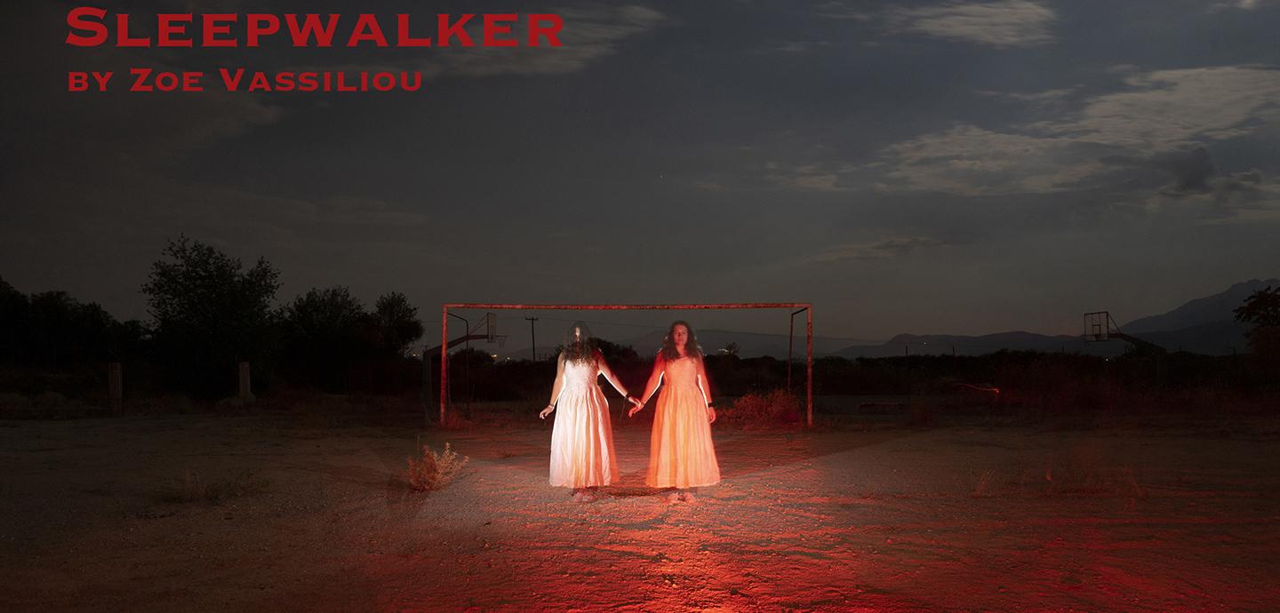 Two figures in long white dresses stand holding hands on red, sandy ground, with the red frame of a swing set behind them. The sky is dark and cloudy, only the two figures are illuminated.