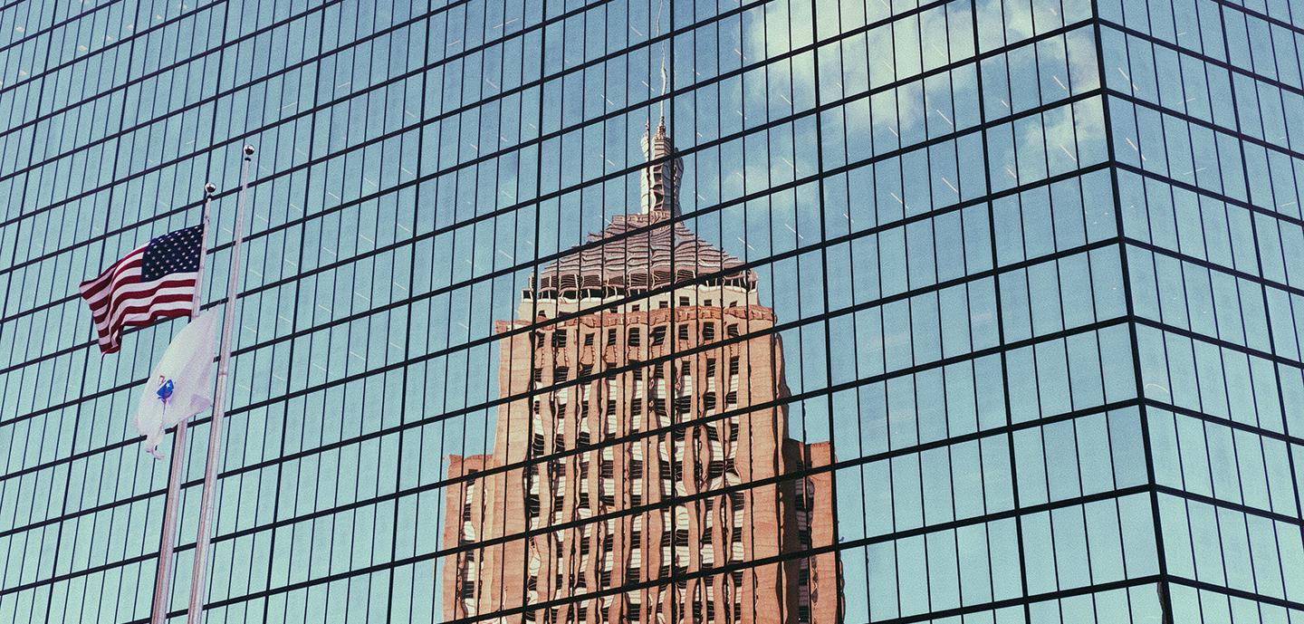 A modern glass building reflects an older skyscraper and the sky, with American flags waving in the foreground.
